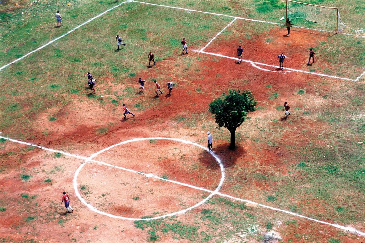 Campo de várzea visto de cima, com uma árvore dentro do gramado e jogadores em volta.